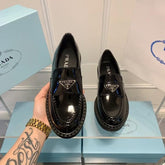 Black Prada loafers held by a hand with a tattoo, on a blue Prada box.